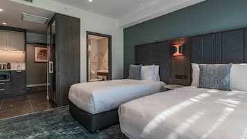 Ett modernt hotellrum med två sängar, en liten sittgrupp, ett litet kök och ett badrum som syns genom en öppen dörr.