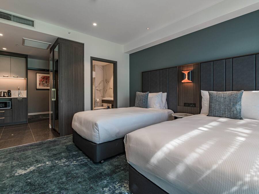 Ett modernt hotellrum med två sängar, en liten sittgrupp, ett litet kök och ett badrum som syns genom en öppen dörr.