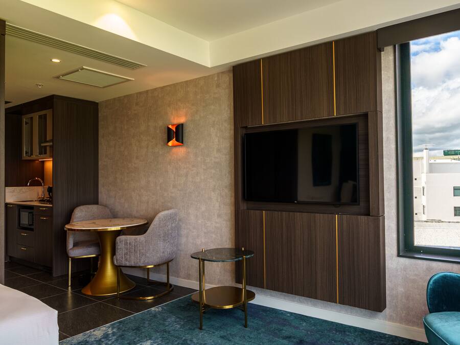 Una habitación de hotel moderna con una televisión montada en la pared, una pequeña mesa redonda, dos sillas, un sofá y vistas a la ciudad a través de la ventana.