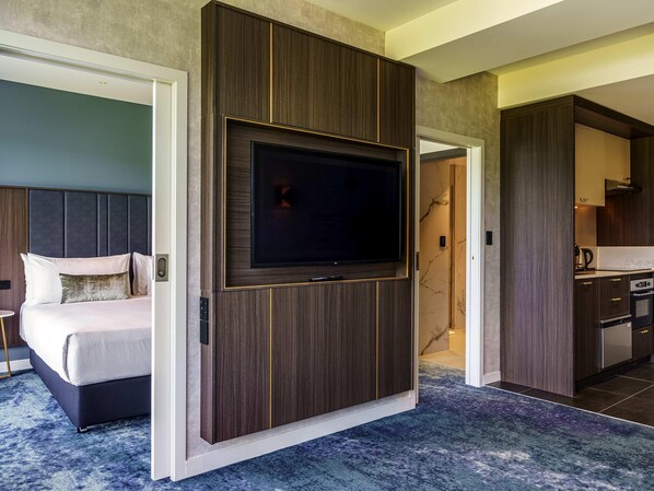A modern hotel room with a large flat-screen TV mounted on a wooden panel wall, a bed with white bedding, a blue carpet, and a separate kitchen area. - The Sebel Wellington Lower Hutt (Lower Hutt)