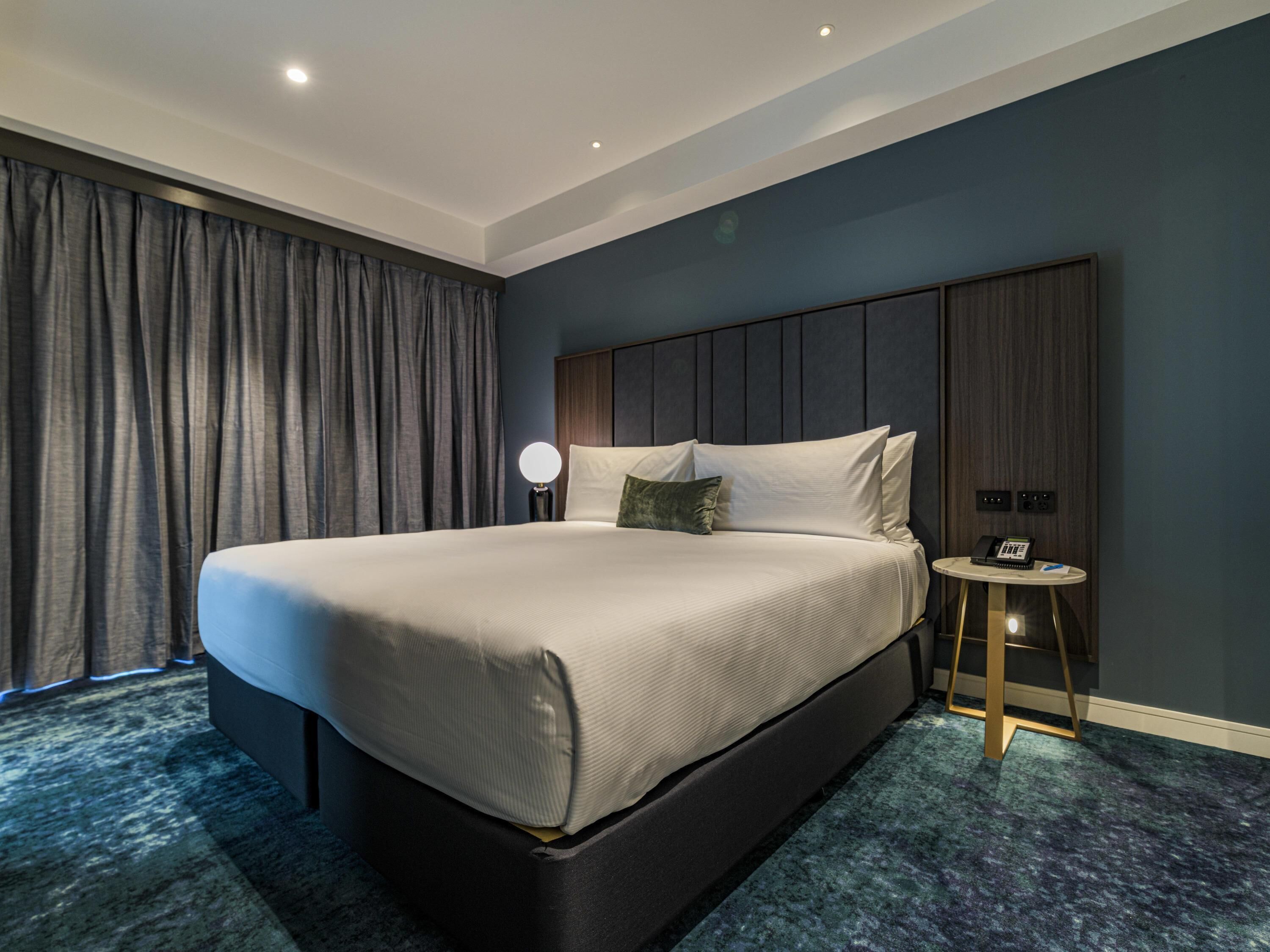 A modern hotel room with a large bed, a bedside table with a lamp, and a small round table with a phone.