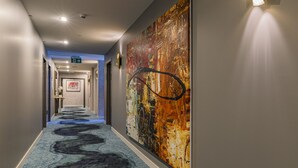 Un couloir d'hôtel avec un tapis bleu, une grande peinture abstraite accrochée au mur et un luminaire fixé au mur.