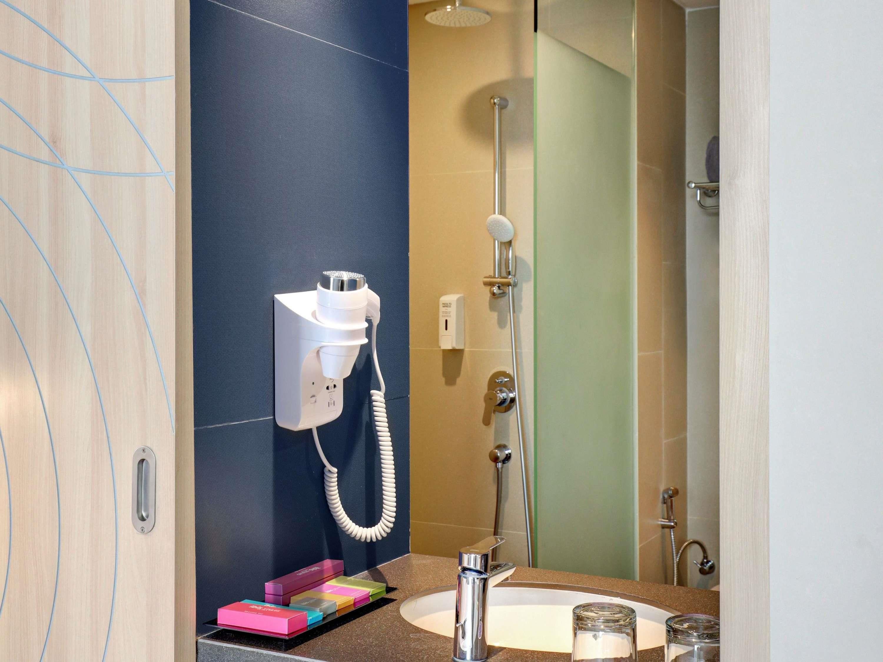 A bathroom with a hairdryer mounted on the wall, a shower, and a sink with a glass jar.