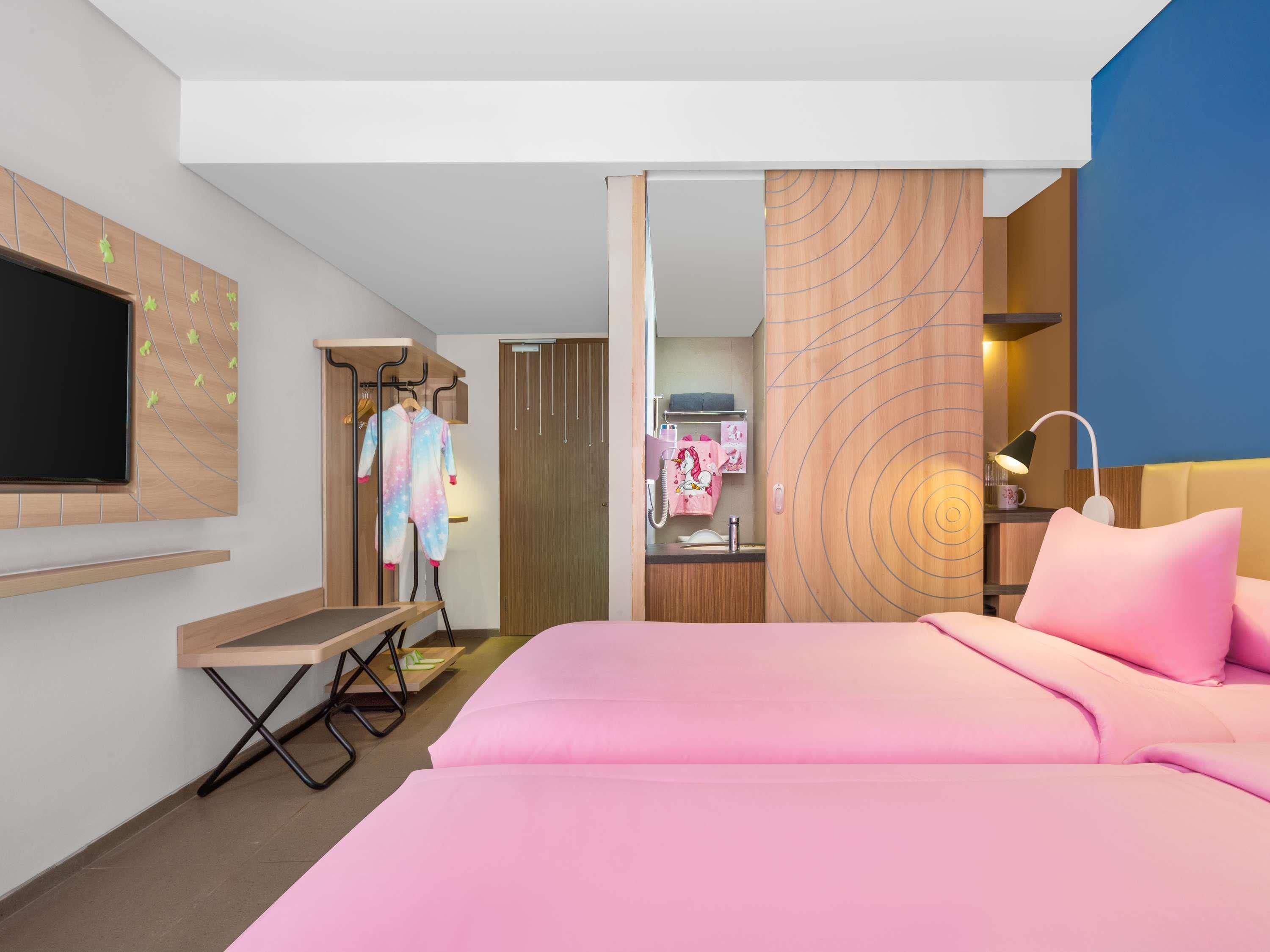 A modern hotel room with a bed, a TV, a desk, and a bathroom visible through an open door.