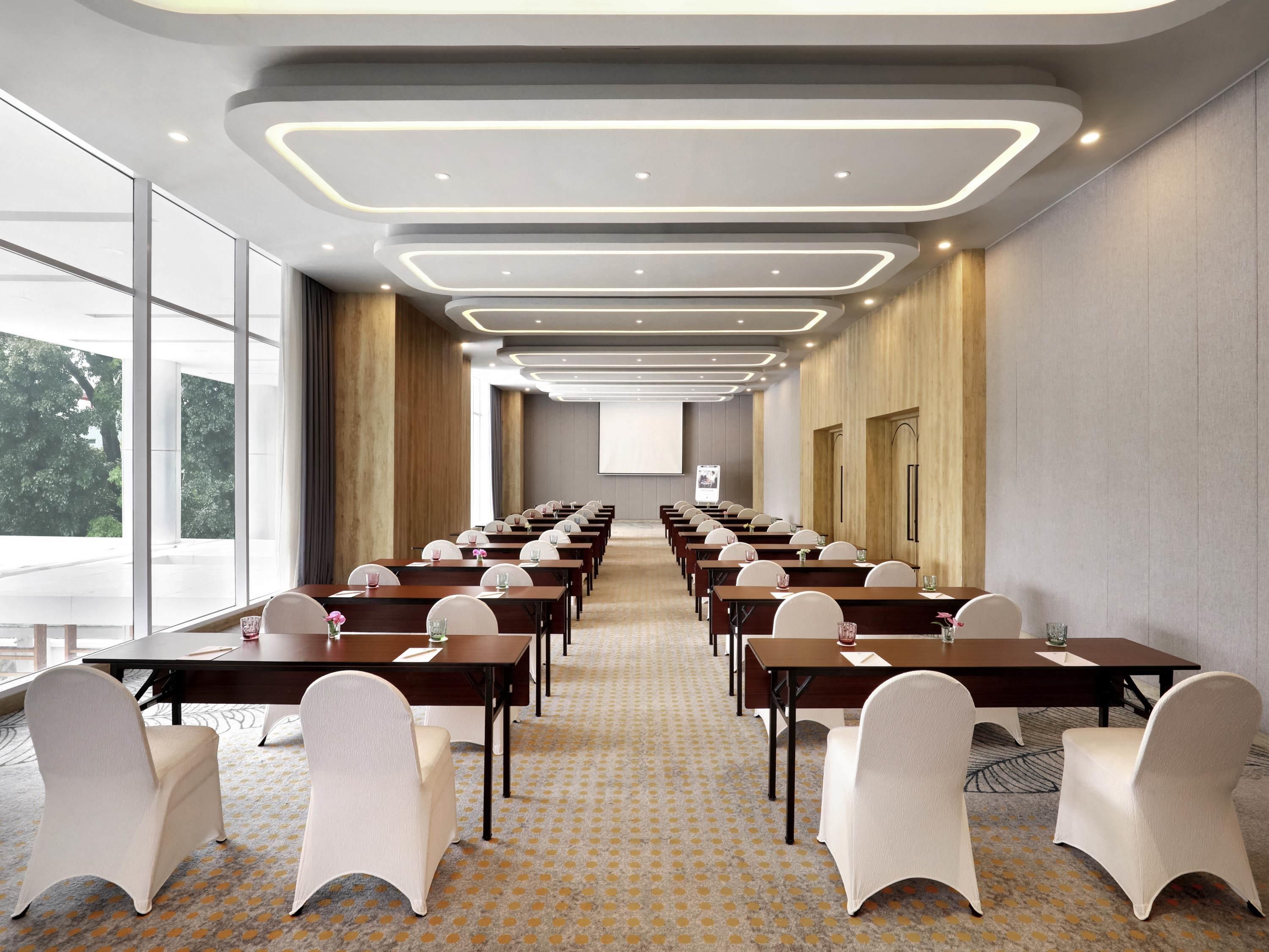 A conference room with rows of tables and chairs, large windows, and a projector screen.