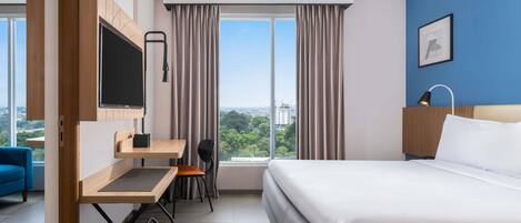A hotel room with a bed, a desk with a chair, a TV, a window with curtains, and a city view.