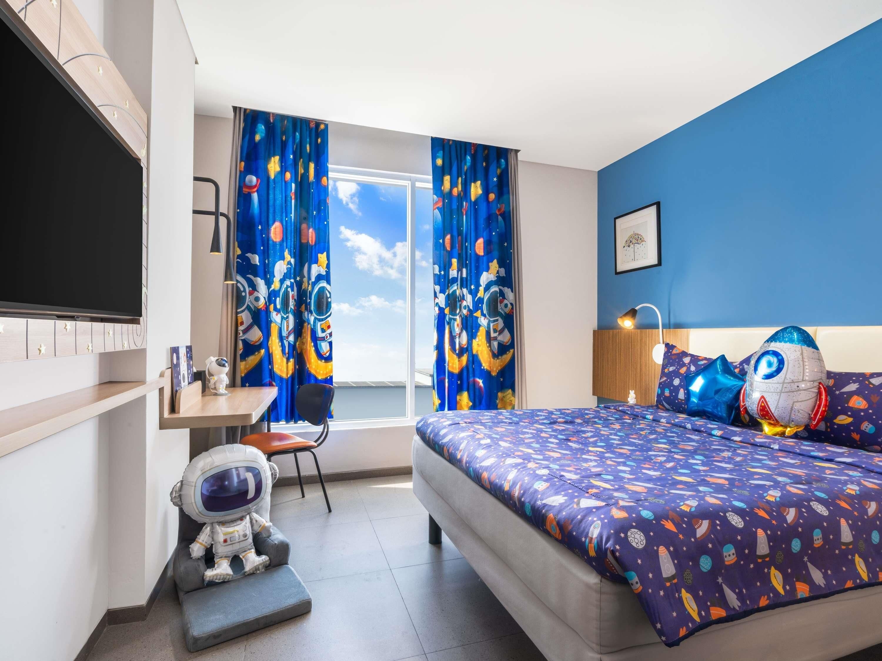 A bedroom with a bed, a desk, a chair, a TV, and a window with curtains.