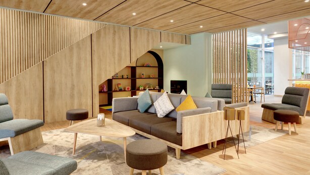 A modern living room with a sofa, ottomans, and a coffee table. There is a staircase leading to a balcony with a view. - ibis Styles Bogor Pajajaran (Bogor)