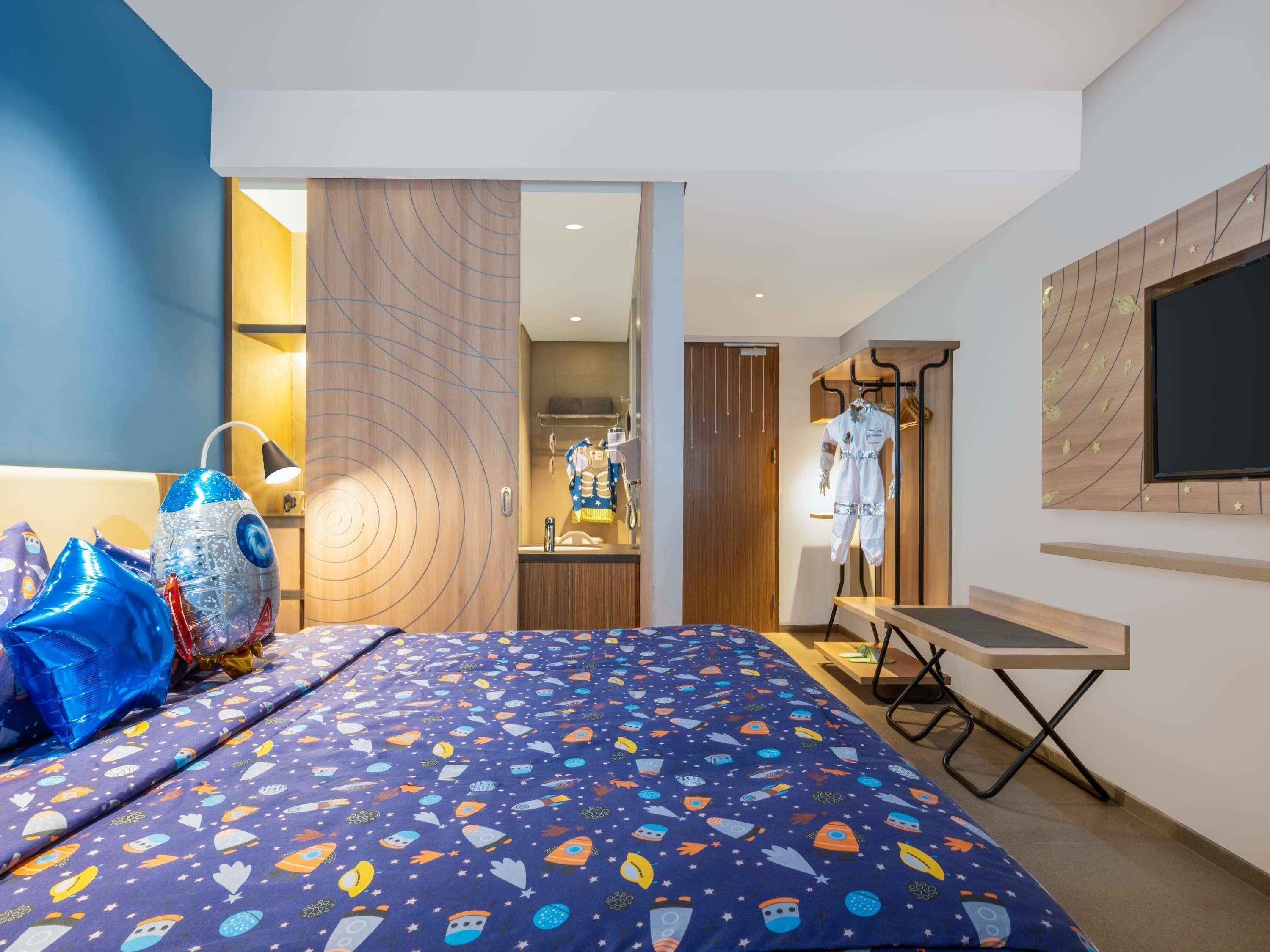 A hotel room with a bed featuring a space-themed bedspread, a wardrobe with a mirror, a desk with a chair, and a TV mounted on the wall.