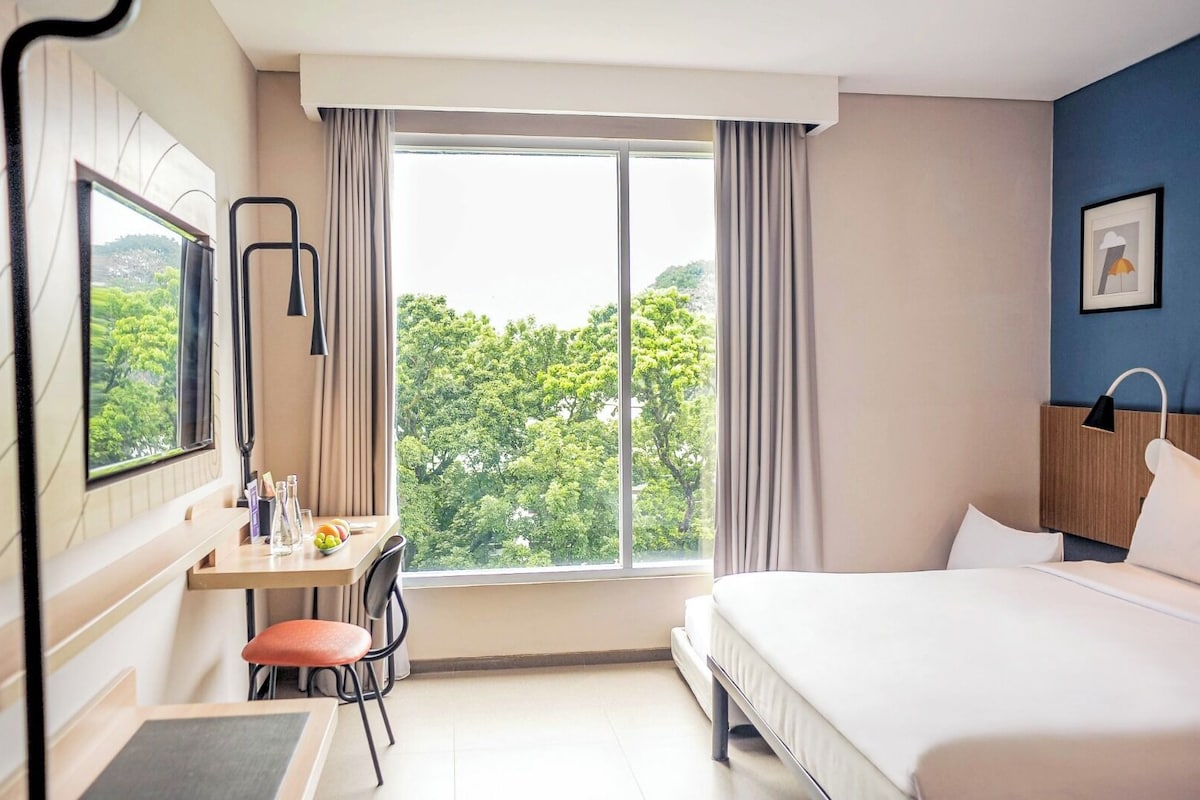 A hotel room with a bed, a desk with a chair, a window with curtains, and a view of trees.