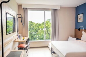 A hotel room with a bed, a desk with a chair, a window with curtains, and a view of trees. - ibis Styles Bogor Pajajaran (Bogor)