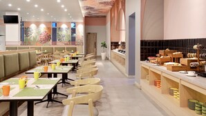 A modern restaurant with long communal tables and booths, a kitchen counter with food items, and a colorful ceiling design. - ibis Styles Bogor Pajajaran (Bogor)