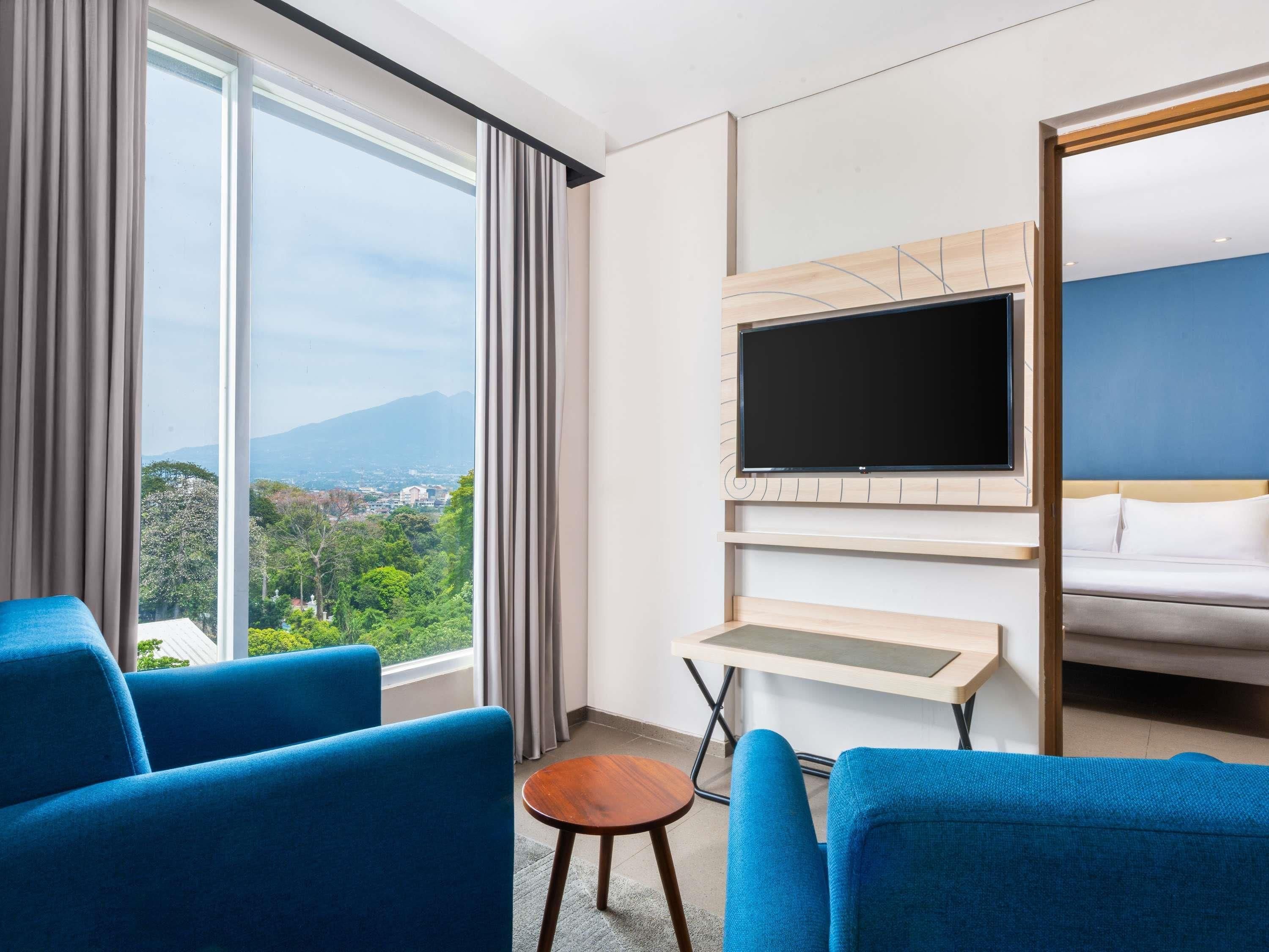 Une chambre d’hôtel dotée d’une grande fenêtre, d’un téléviseur à écran plat fixé au mur, d’un lit et de deux fauteuils bleus.