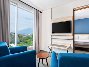 Habitación de hotel con un ventanal grande, un televisor de pantalla plana montado en la pared, una cama y dos sillones azules.