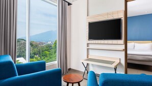 A hotel room with a large window, a flat-screen TV mounted on the wall, a bed, and two blue armchairs. - ibis Styles Bogor Pajajaran (Bogor)