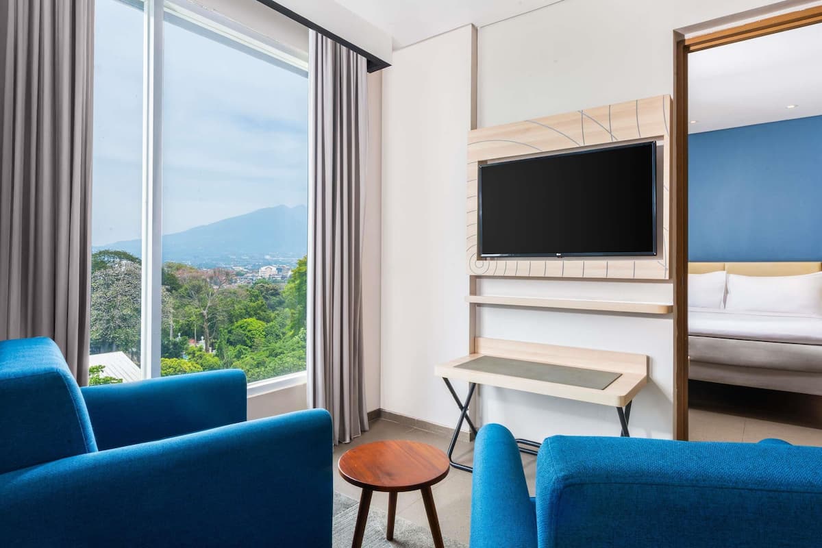 A hotel room with a large window, a flat-screen TV mounted on the wall, a bed, and two blue armchairs.