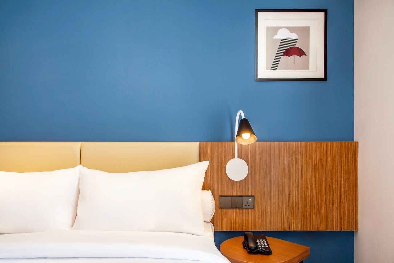 A hotel room with a bed, a bedside table with a lamp, a framed picture on the wall, and a wooden headboard.