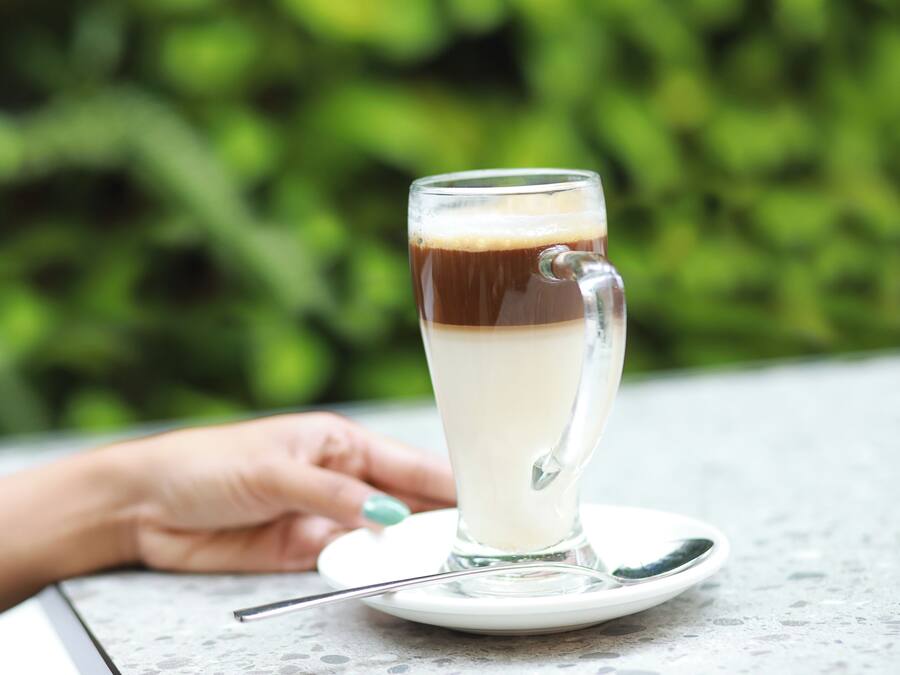 A tall glass of layered beverage on a saucer with a spoon, placed on a table with a blurred green background.