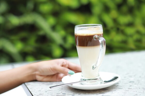 A tall glass of layered beverage on a saucer with a spoon, placed on a table with a blurred green background. - ibis Styles Bogor Pajajaran (Bogor)