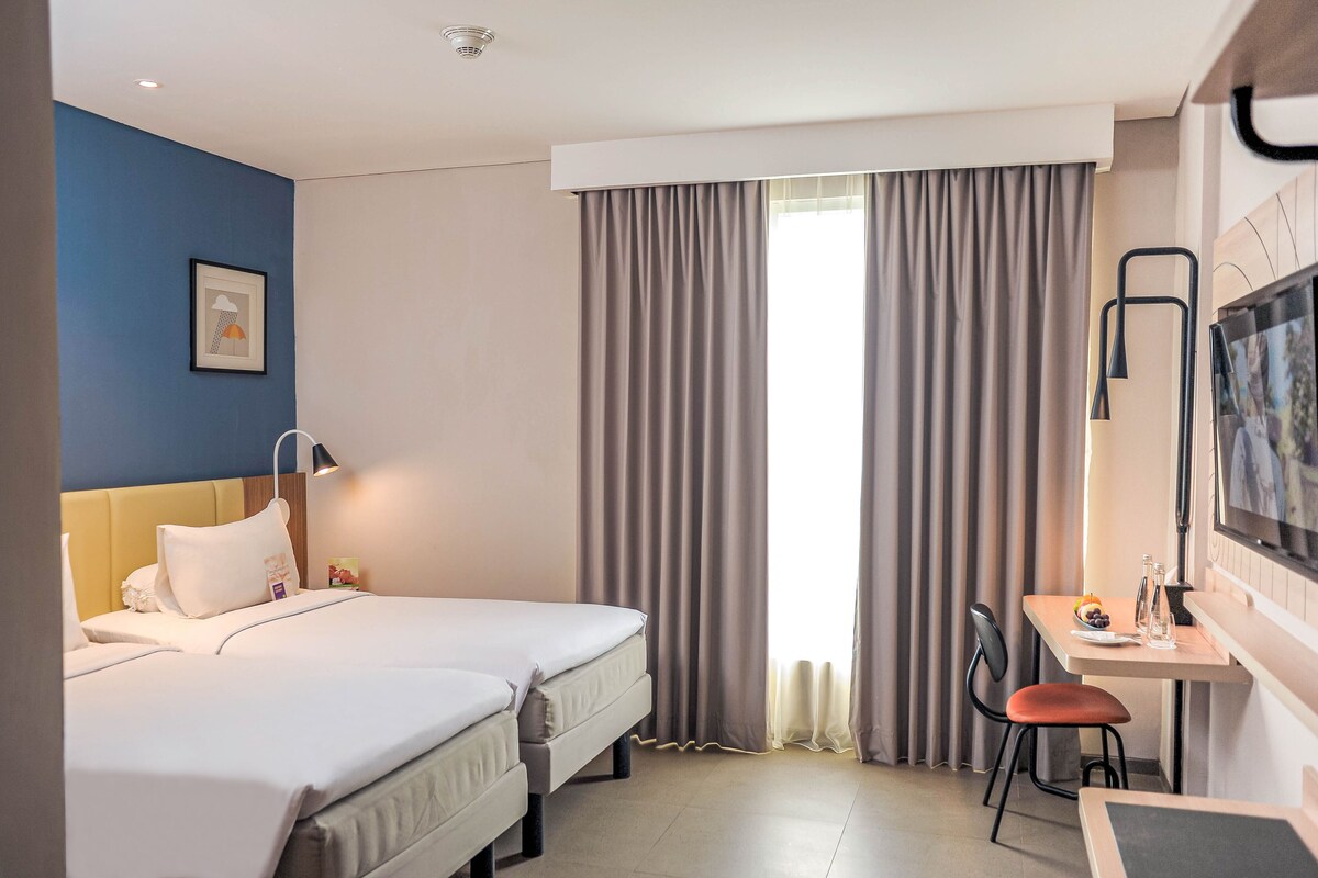 A hotel room with two beds, a desk with a chair, a TV, and a window with curtains.