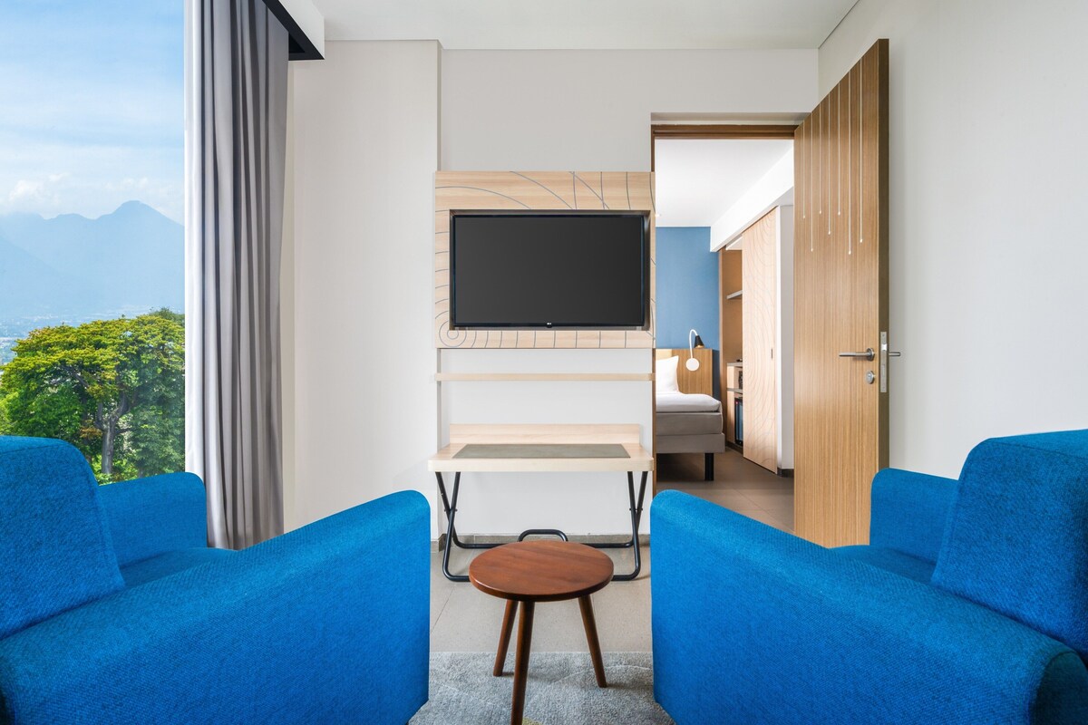 A modern hotel room with a TV mounted on a wall, a bed, a desk, and two blue armchairs.