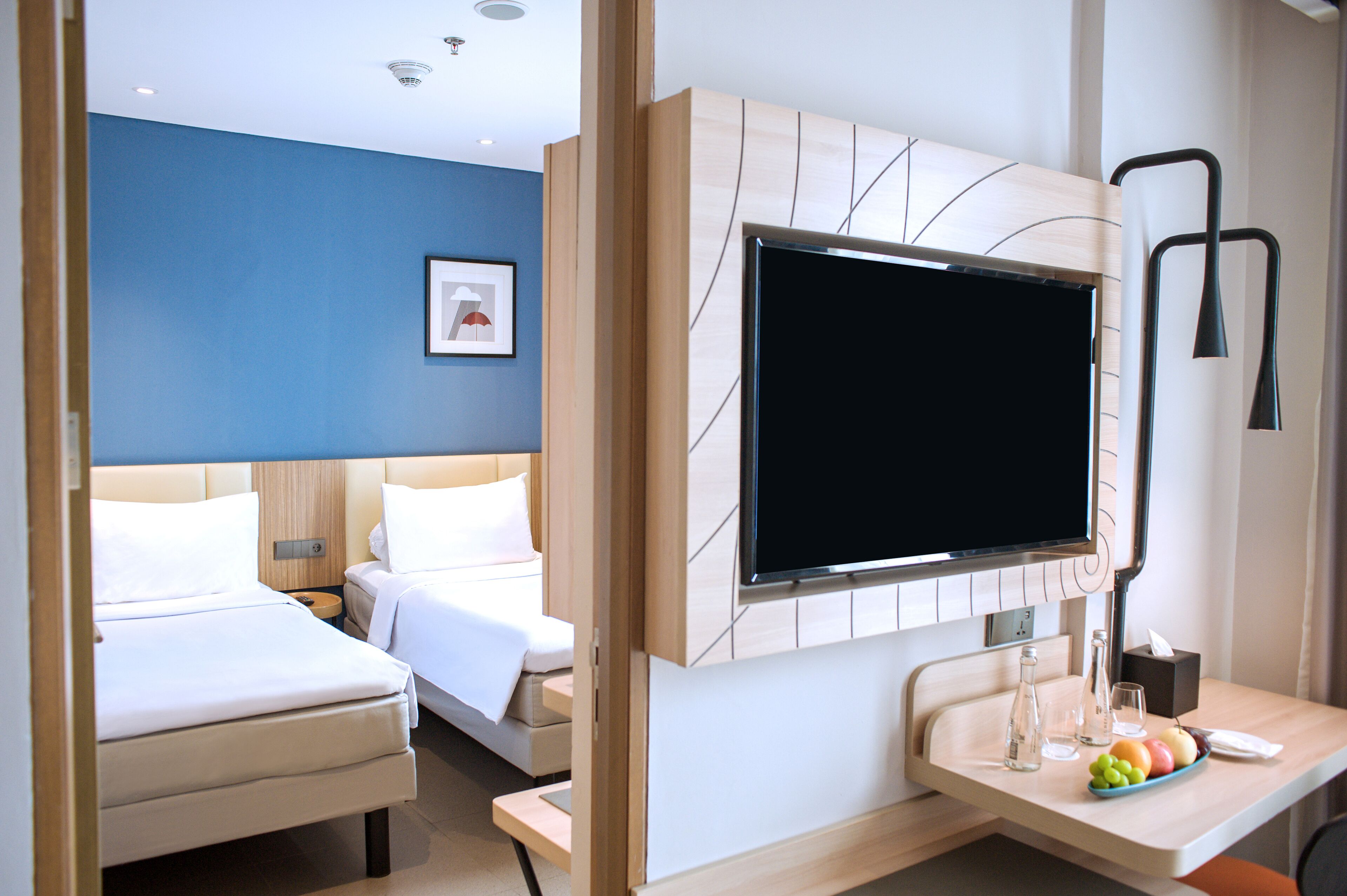 Habitación de hotel con dos camas, televisión, escritorio con frutero y mesita de noche con lámpara.