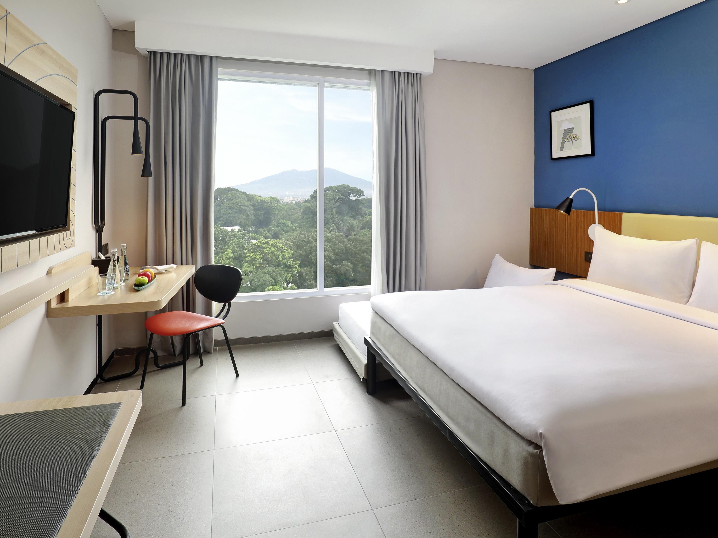 Hotelkamer met een bed, bureau, stoel en een groot raam met uitzicht op groen en bergen.