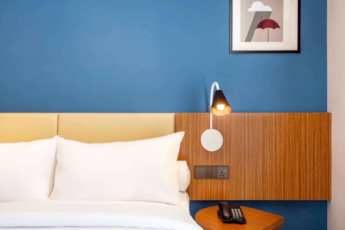 A hotel room with a bed, a bedside table with a telephone, a wall-mounted light, and a framed picture on the wall.
