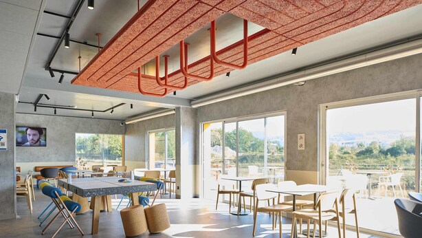 Un espace intérieur moderne, doté de tables et de chaises, de grandes fenêtres et d’un plafond agrémenté d’éléments structurels rouges.