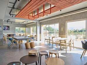 Un espace intérieur moderne, doté de tables et de chaises, de grandes fenêtres et d’un plafond agrémenté d’éléments structurels rouges.