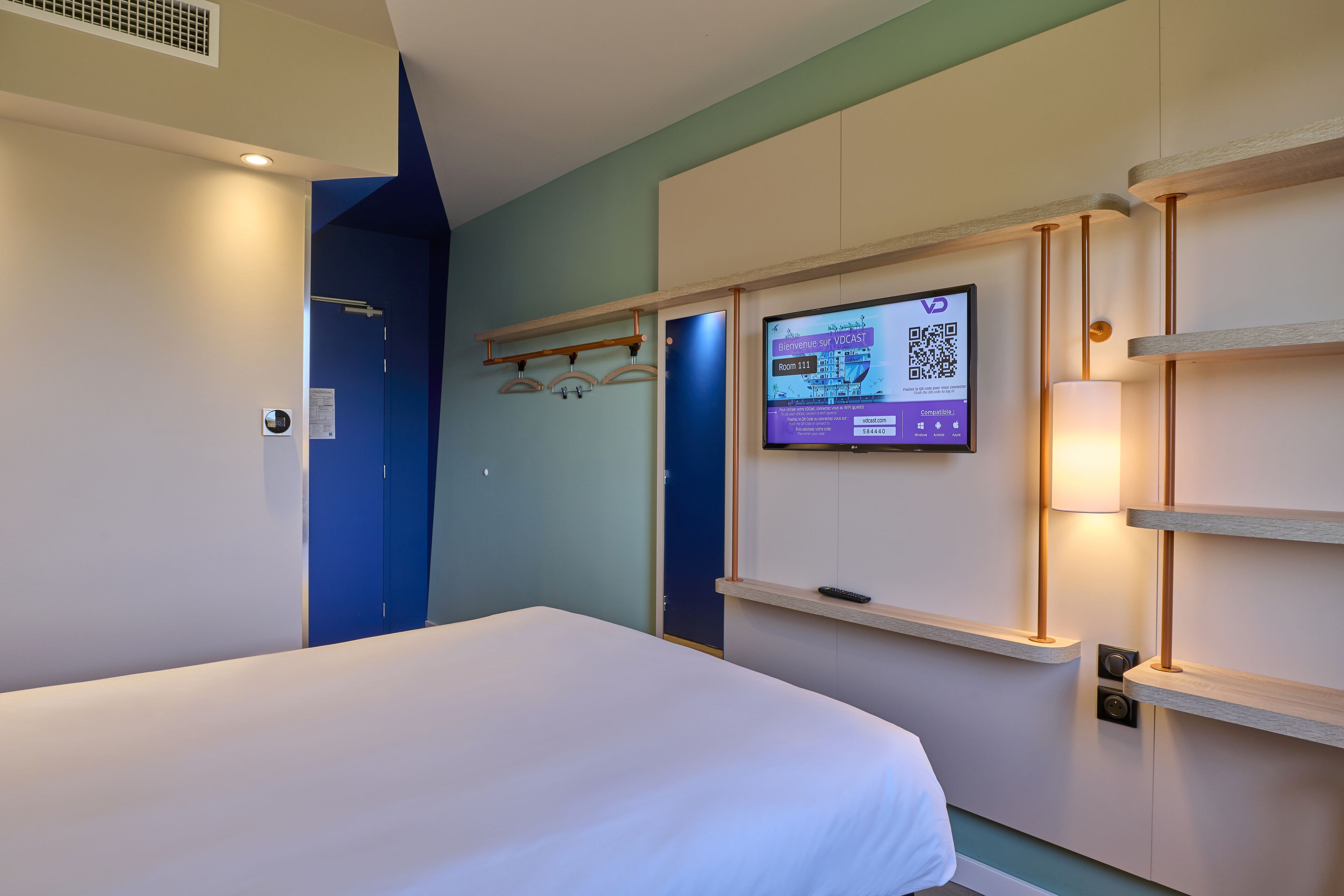 A hotel room with a bed, a TV mounted on the wall, and a door with a blue frame.