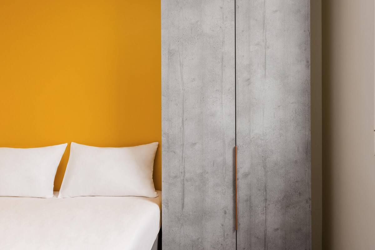 A bed with white bedding, a concrete wall, and a yellow wall.