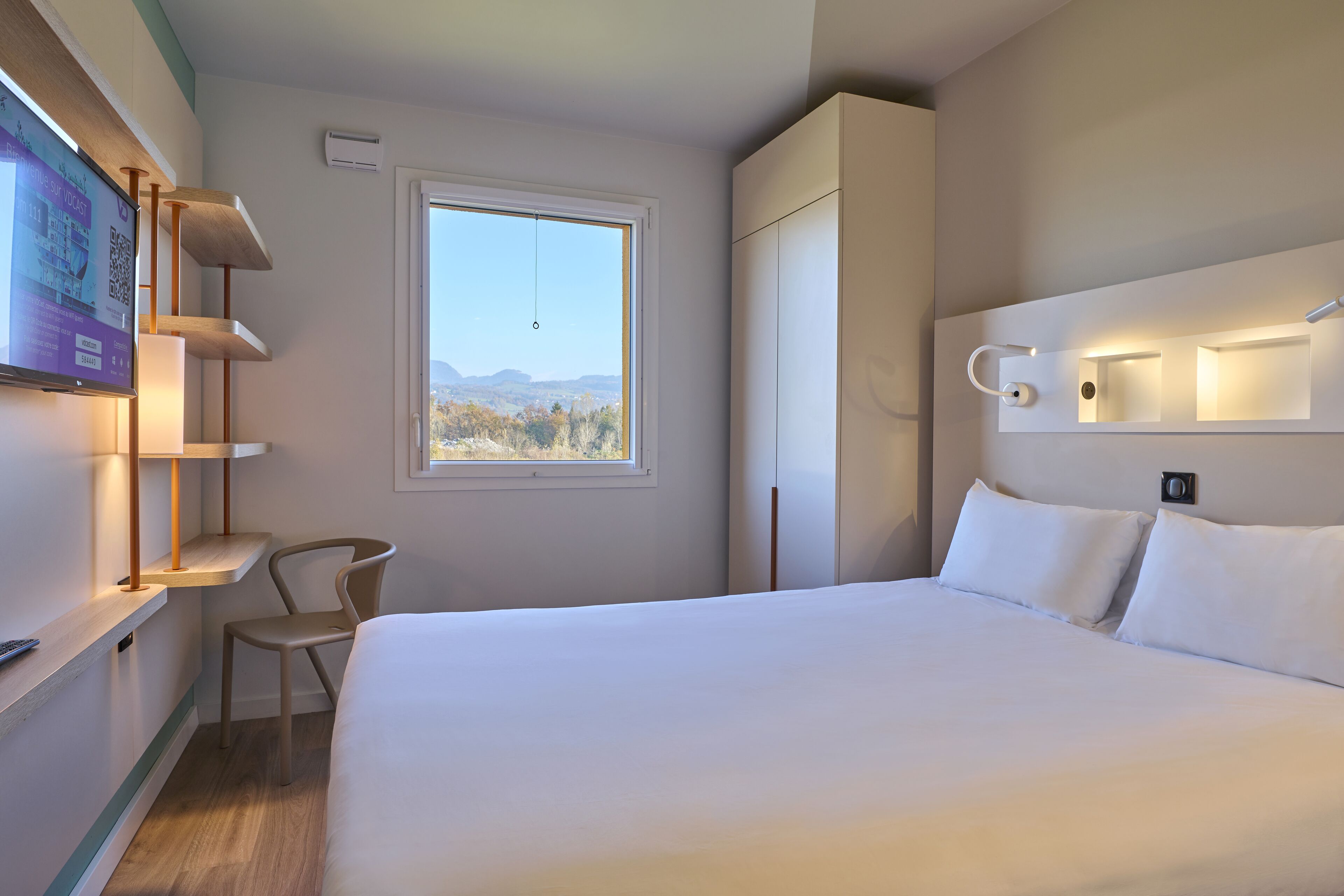 Habitación de hotel moderna con una cama grande, un escritorio con silla, un televisor y una ventana con vistas a las montañas.