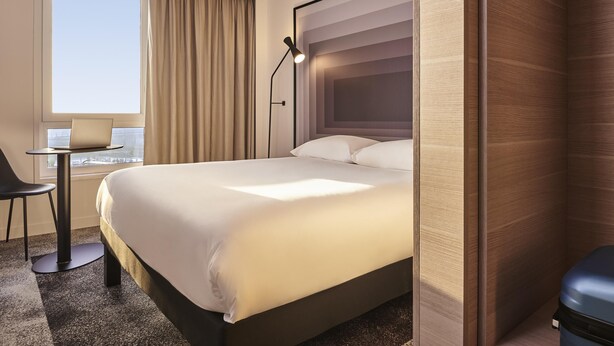 A hotel room with a bed, a desk with a chair, a laptop, a window with curtains, and a view of the city. - ibis Bethune Centre Gare (Béthune)
