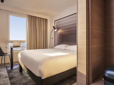 A hotel room with a bed, a desk with a chair, a laptop, a window with curtains, and a view of the city.. ibis Bethune Centre Gare