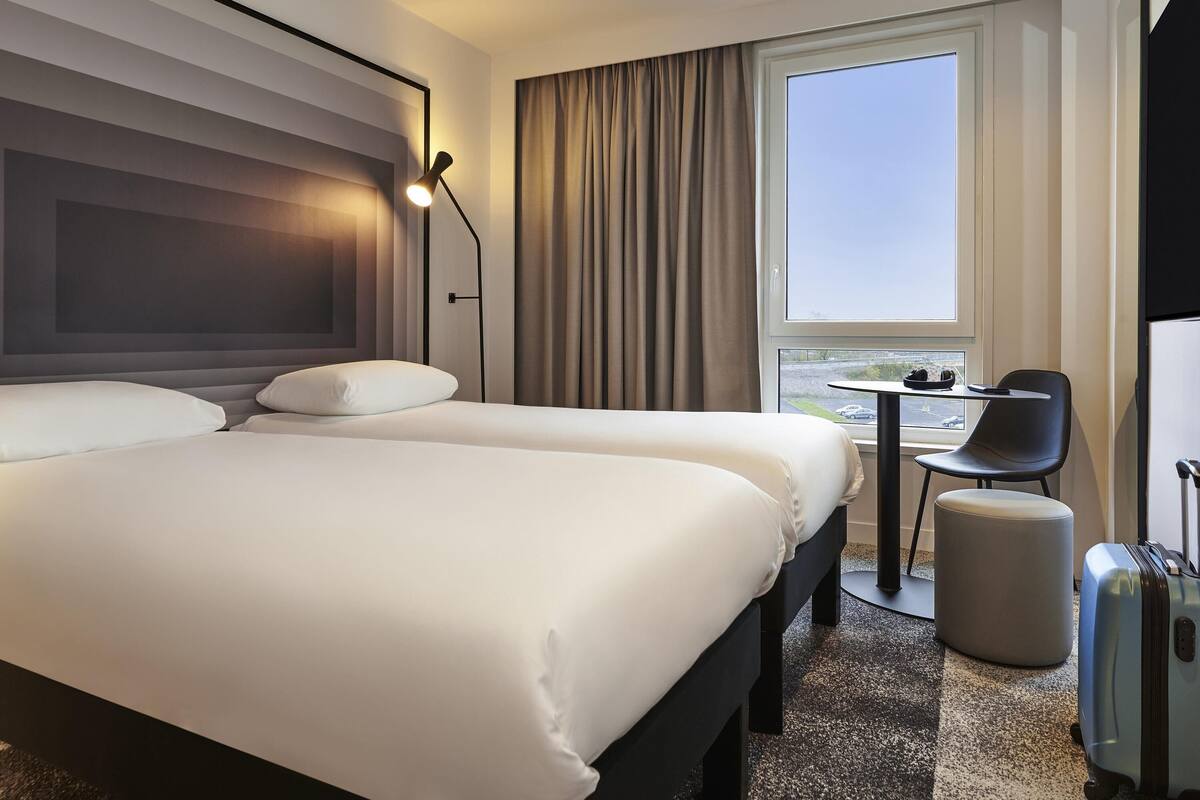 Une chambre d’hôtel avec un grand lit, une petite table avec une chaise, une valise et une fenêtre donnant sur un beau paysage.