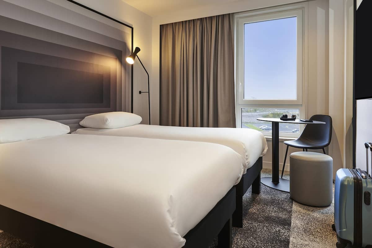 Une chambre d’hôtel avec un grand lit, une petite table avec une chaise, une valise et une fenêtre donnant sur un beau paysage.