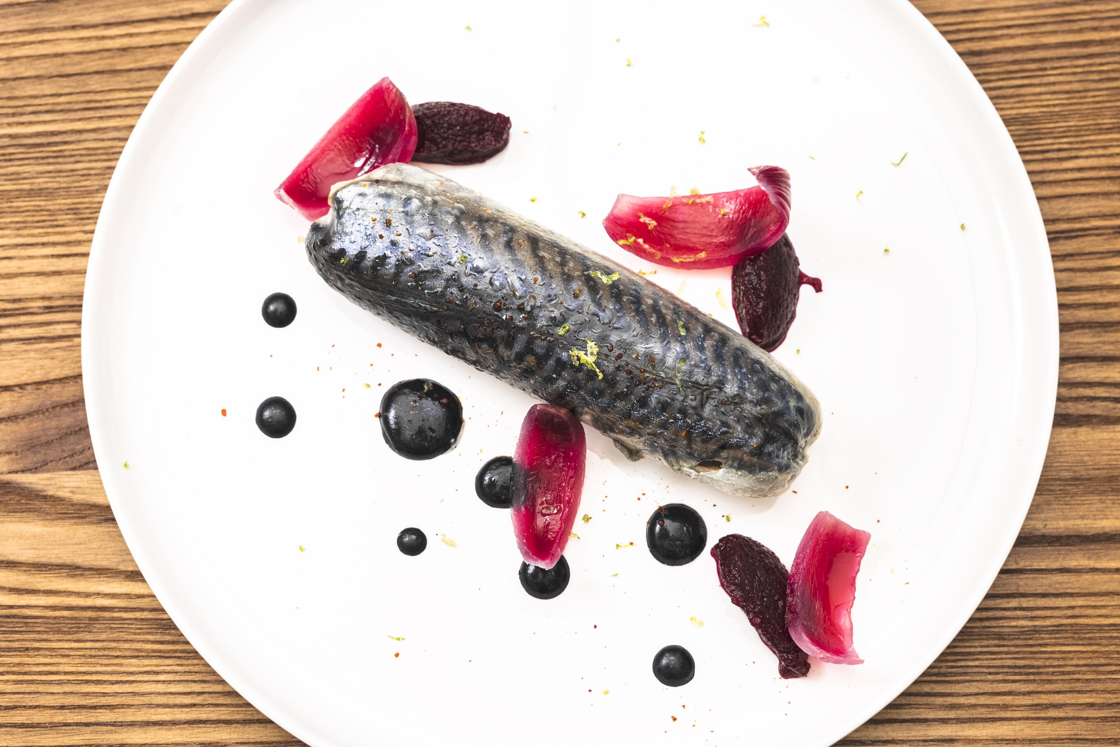 A white plate with a fish fillet, black and red garnishes, and a wooden table background.