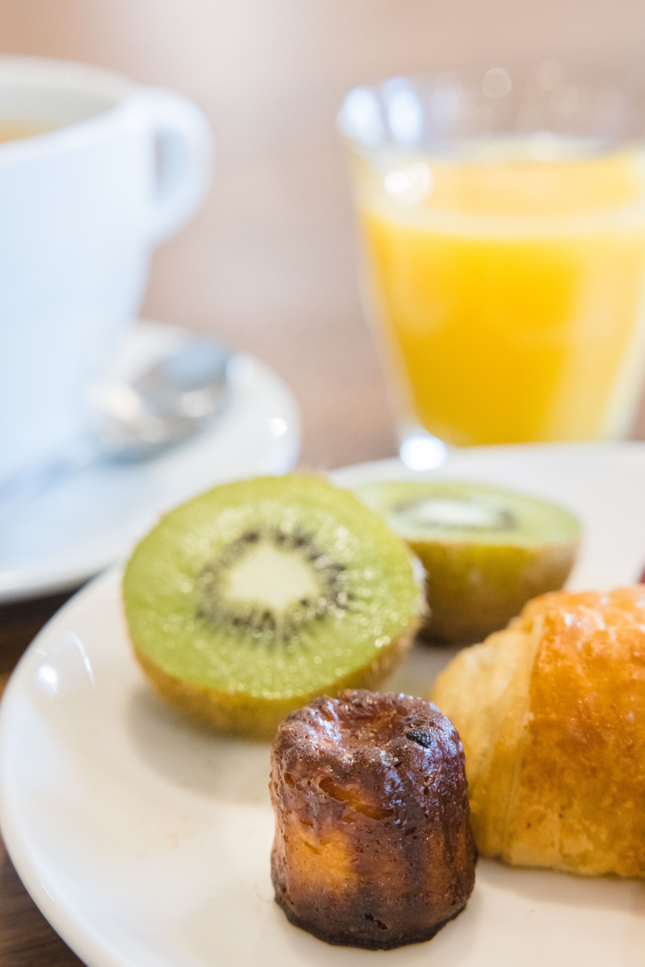 Un plato con un croissant, una rodaja de kiwi y un muffin, junto a una taza y un vaso de jugo de naranja.