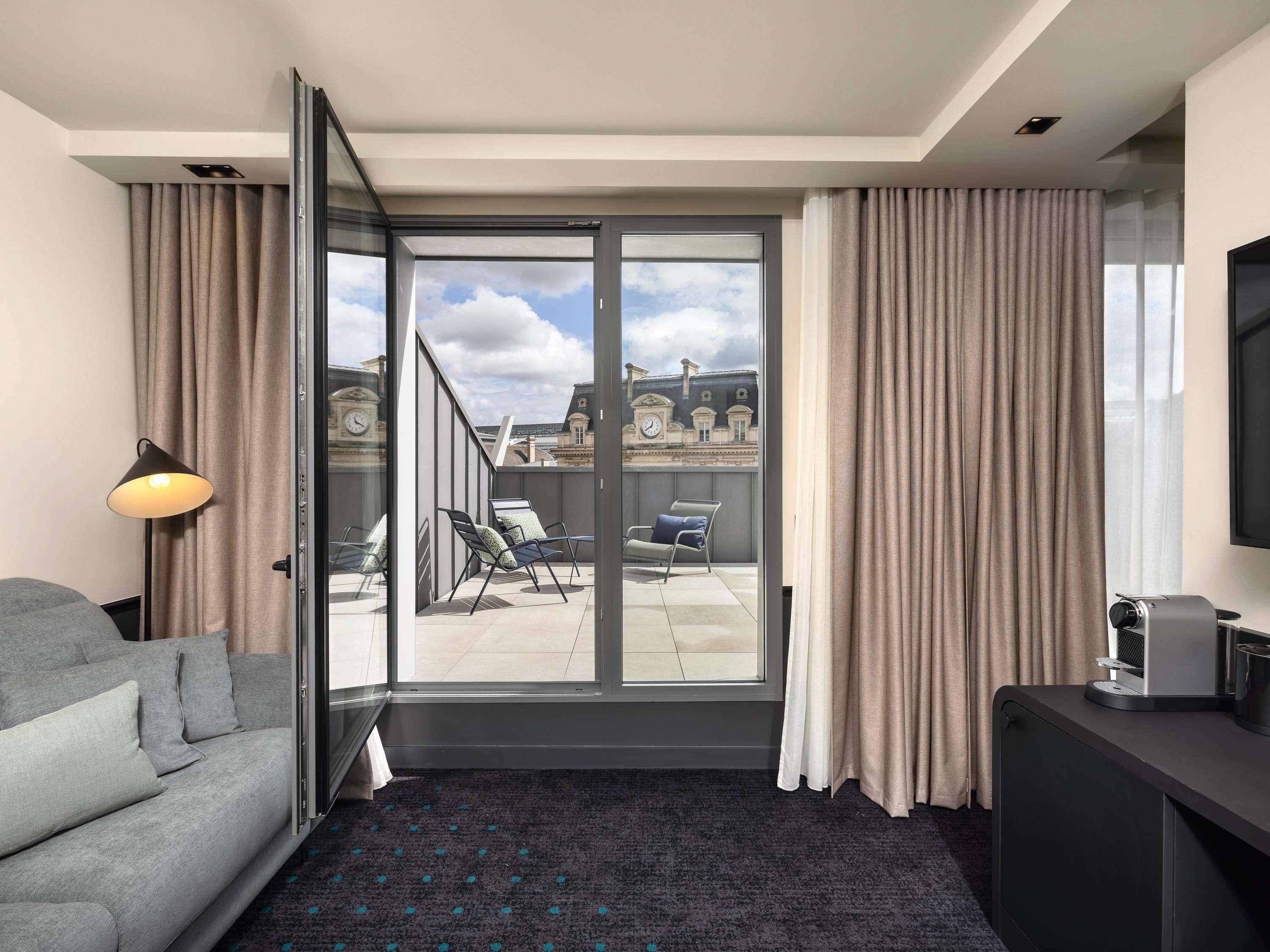 A modern hotel room with a grey sofa, a small table with a coffee machine, a balcony with a view of historic buildings, and a sliding glass door.
