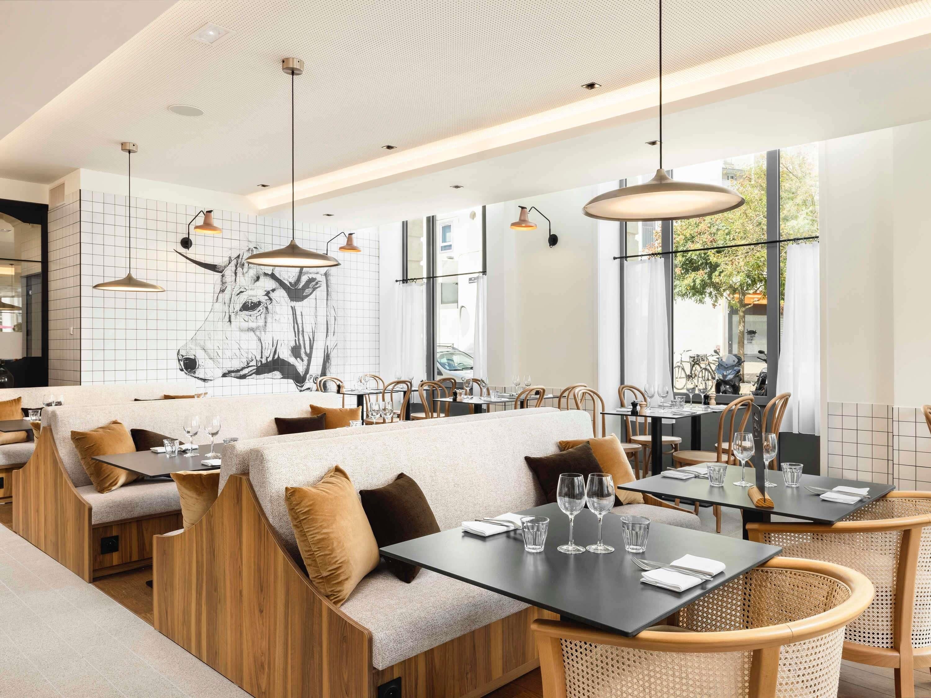 Un intérieur de restaurant moderne, avec de hauts plafonds, des luminaires suspendus et une grande fresque murale.