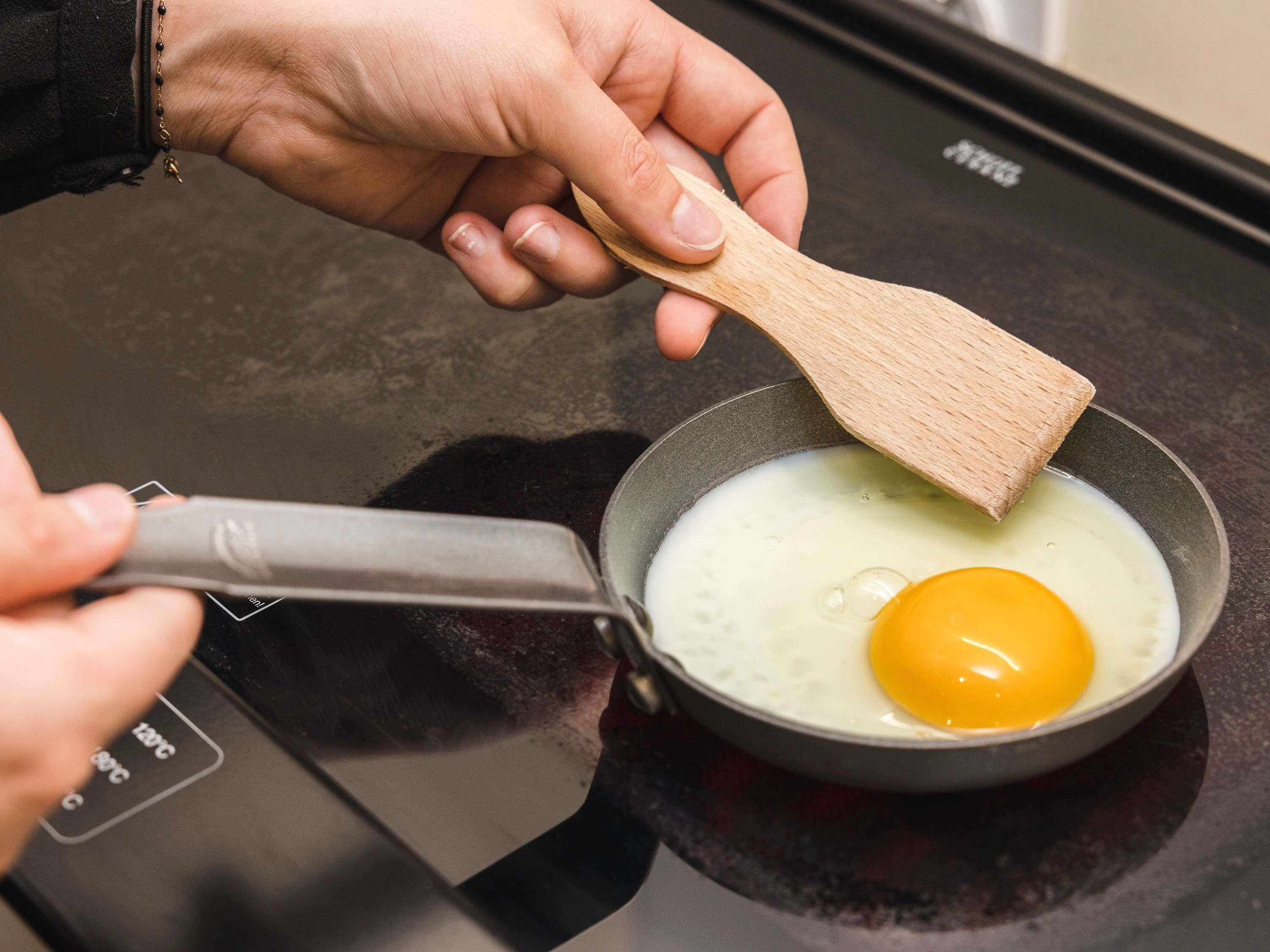 A hand holding a wooden spatula over a frying pan with an egg cooking on an induction cooktop.