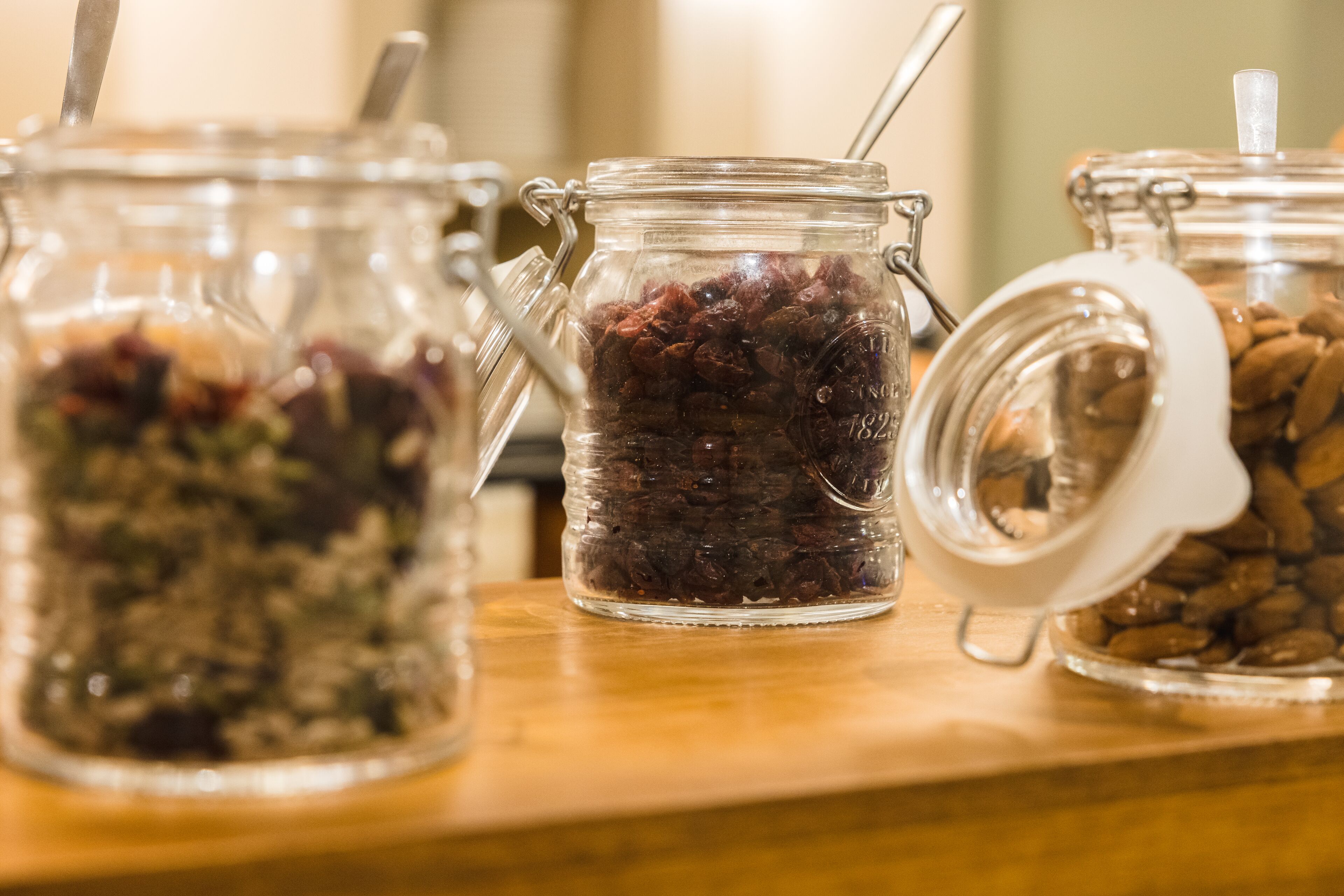 Three glass jars with metal handles containing various dried fruits and nuts on a wooden surface.