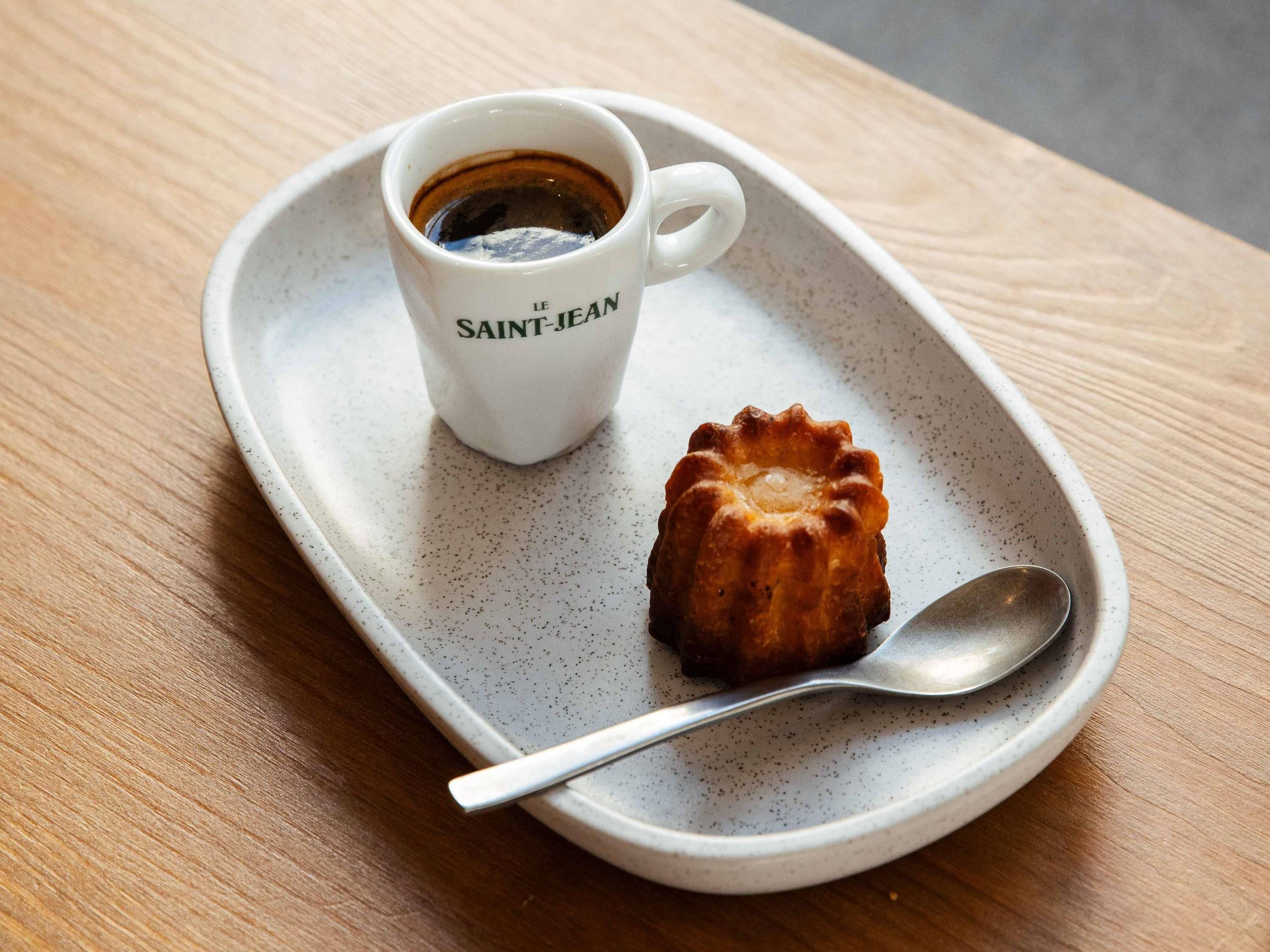 A white ceramic cup labeled "SAINT- JEAN" with a dark liquid, possibly coffee, and a small pastry on a speckled ceramic plate with a spoon.