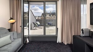 Een moderne hotelkamer met een grijze bank, een kleine tafel met een koffieapparaat, een balkon met uitzicht op historische gebouwen en een schuifglazen deur.