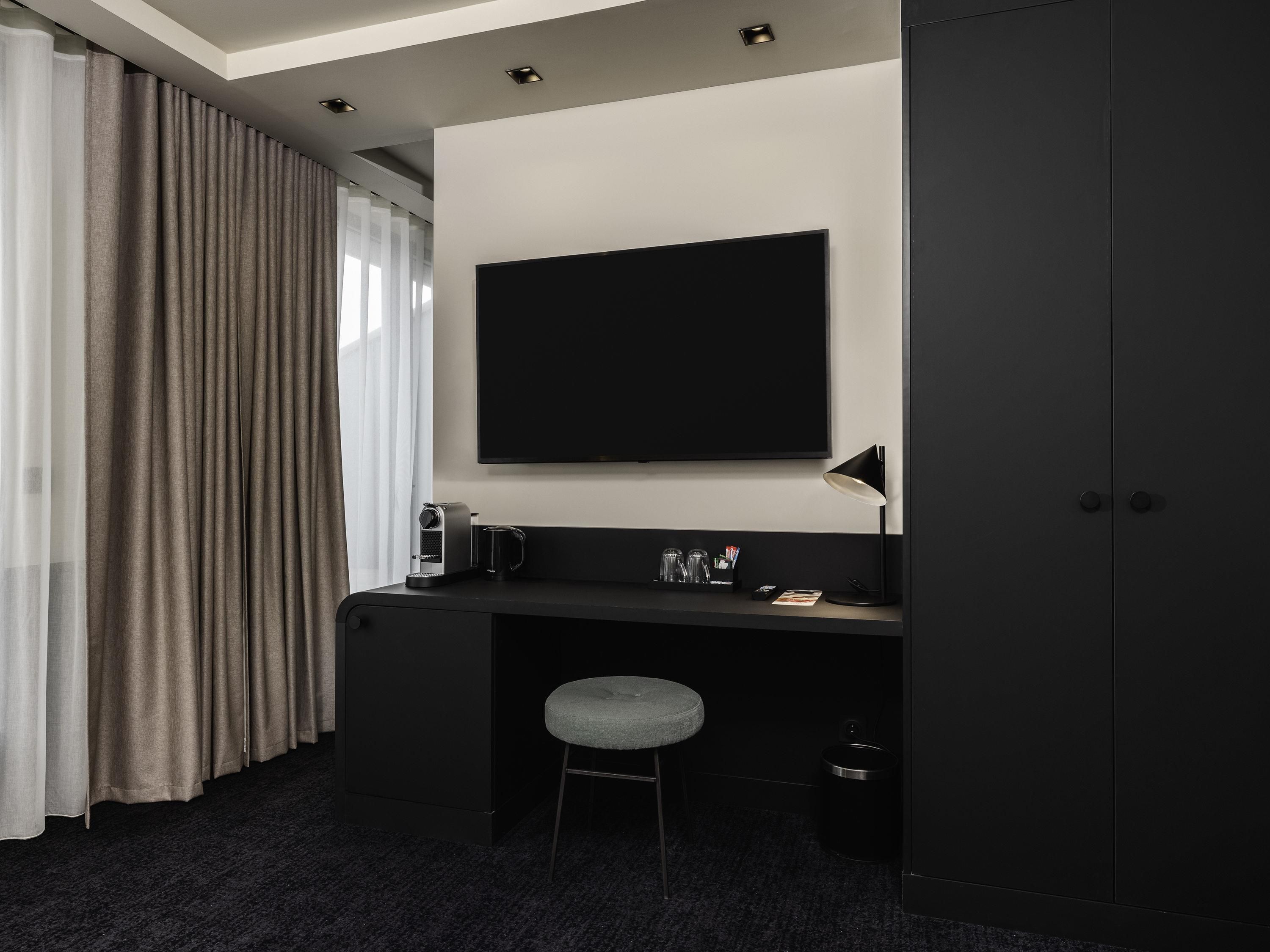 A modern hotel room with a dark desk, a flat-screen TV, a small stool, and a wardrobe.