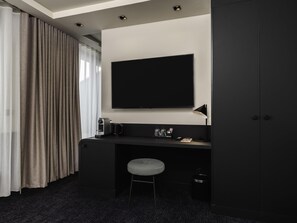 Quarto de hotel moderno com escrivaninha escura, TV de tela plana, um banquinho pequeno e um guarda-roupa.
