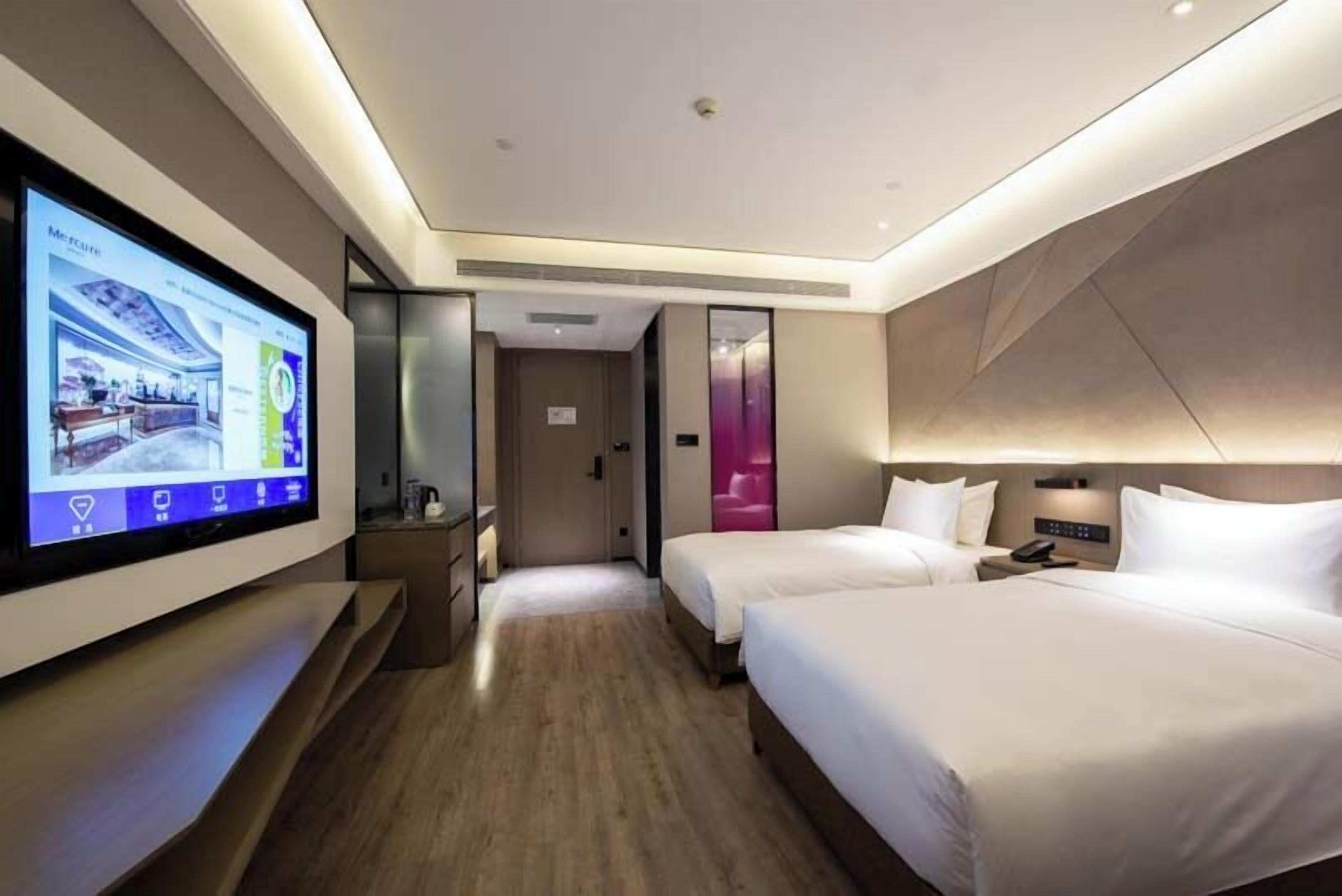 A hotel room with a large TV on the wall, two beds, a bedside table, and a door leading to another room.