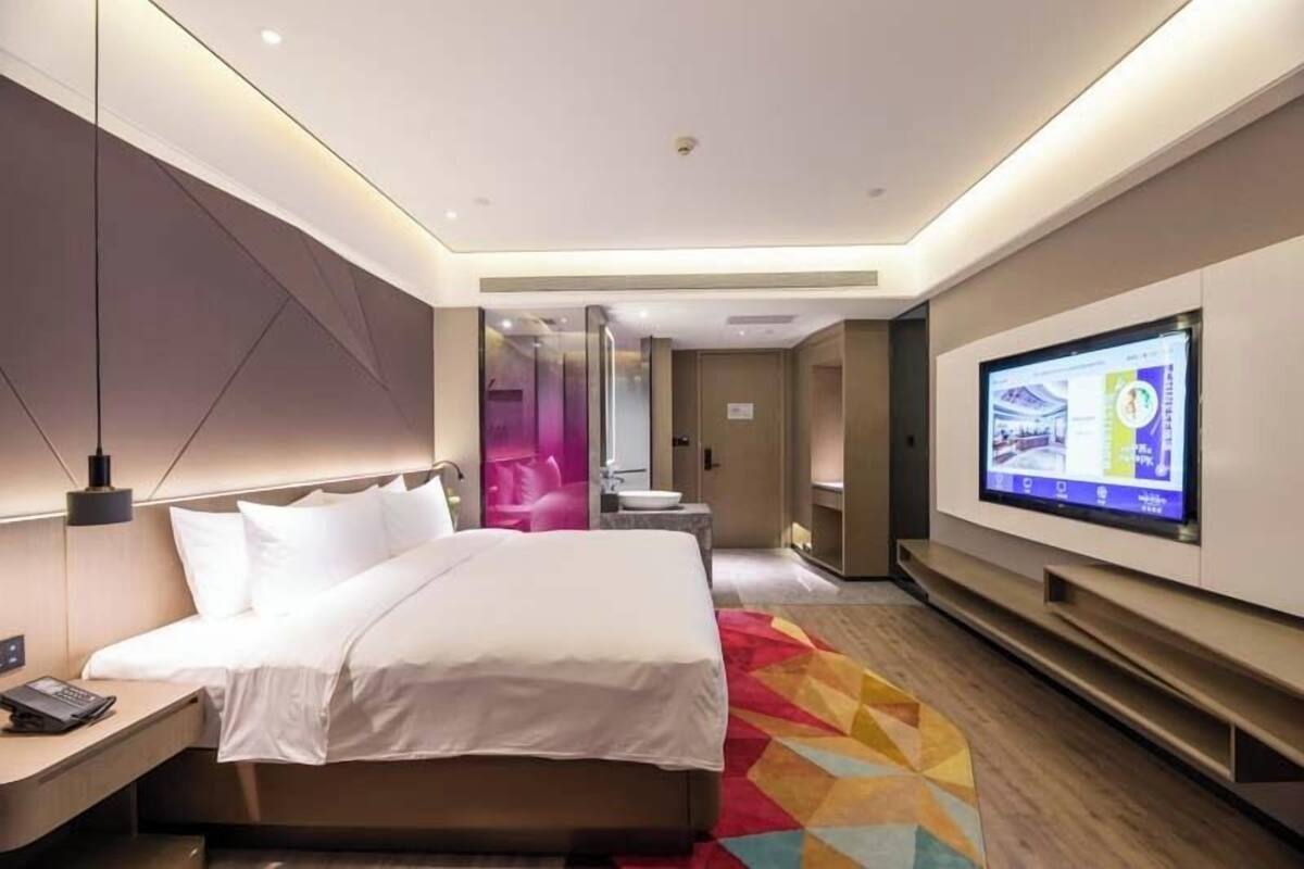 A modern hotel room with a bed, bedside table, TV, and a colorful rug.