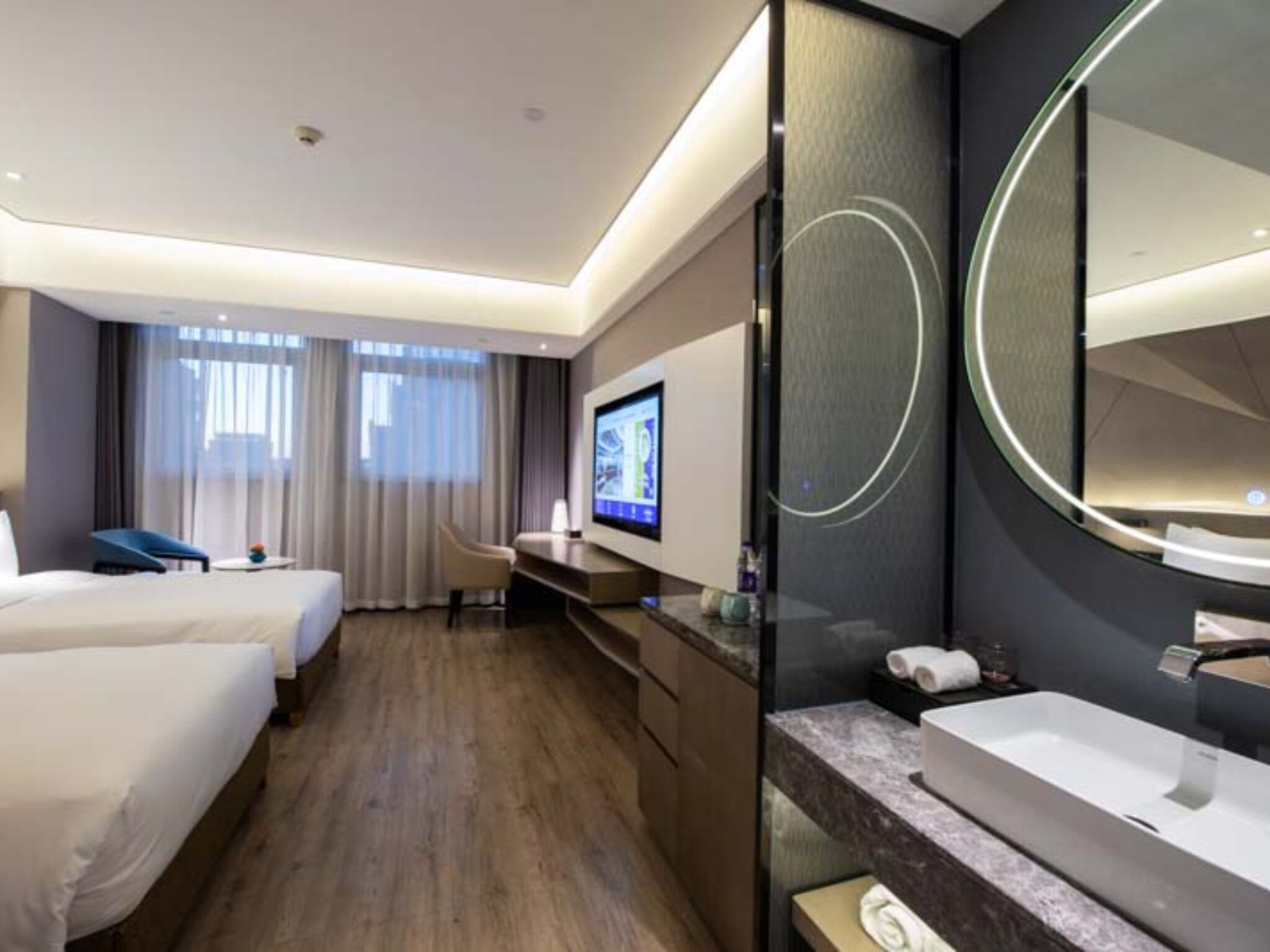 Camera d'albergo con due letti, televisore, scrivania, uno specchio grande e bagno con lavandino e specchio.